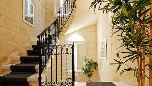A staircase with black railings leading to an upper floor, surrounded by beige stone walls and a large bamboo plant. - Casa Maltija Sliema 4 Bedroom (Sliema)