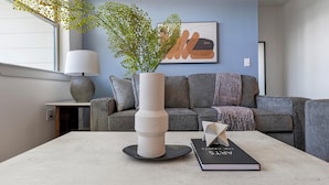Una sala de estar moderna con un sofá gris, un jarrón blanco con follaje verde, un libro negro titulado "ARTS" y una lámpara sobre una mesita.
