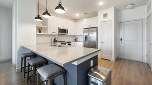 A modern kitchen with white cabinetry, a large island with bar stools, stainless steel appliances, and pendant lighting. - Landing | Chic 2BD, Gym, Clubhouse (Austin)