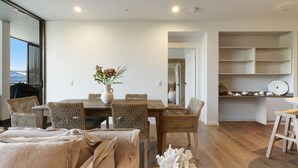 Uma sala de jantar com mesa de madeira e cadeiras de fibra, um vaso com flores e vista para o exterior pela janela.