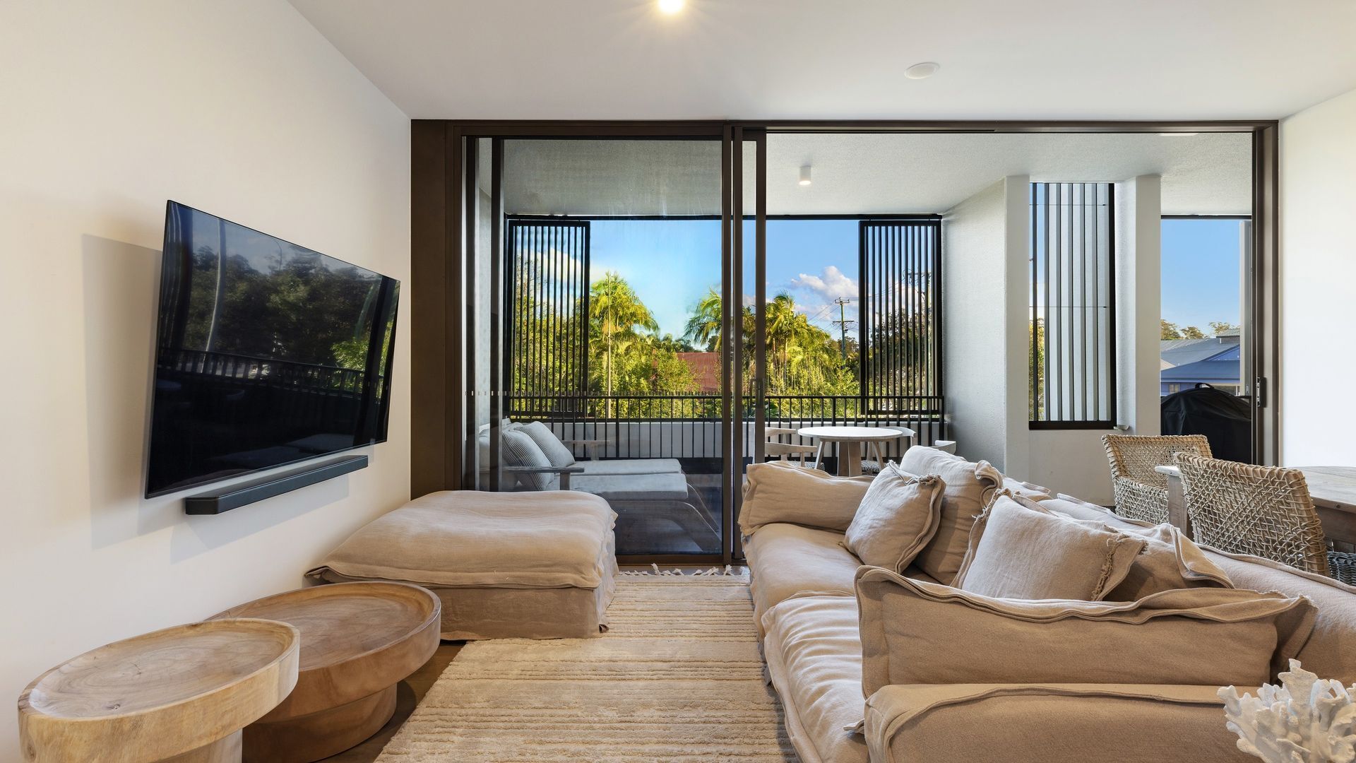 A modern living room with a flat-screen TV, a sofa, a coffee table, and a view of the outdoors through large windows.