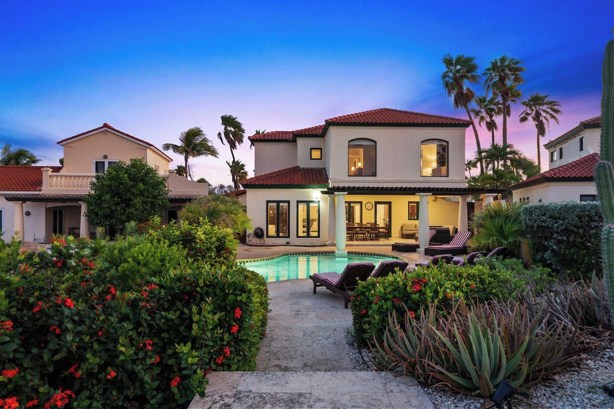 A two-story house with a swimming pool, surrounded by lush greenery and palm trees.