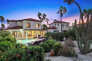 Une maison à deux étages avec une piscine, entourée de cactus et de palmiers.