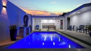 A modern outdoor pool area with a bench and seating, surrounded by a white building with a sunset view. - Private pool • 5 min drive to beach • BBQ (Noord)