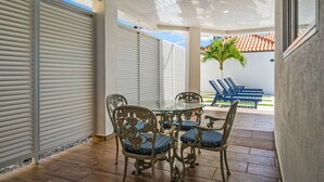 A covered patio area with a table and chairs, a palm tree, and a view of a tiled roof. - Private pool • 5 min drive to beach • BBQ (Noord)