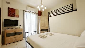 Une chambre à coucher avec un lit, un téléviseur posé sur une commode en bois, un luminaire au plafond et une fenêtre avec des rideaux.