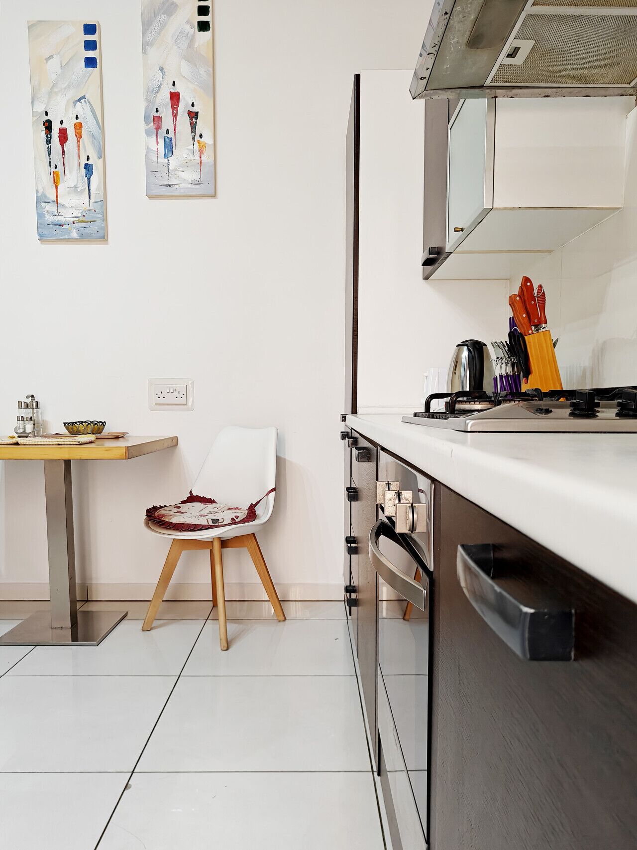 Een moderne keuken met een wit aanrecht, roestvrijstalen apparatuur en een stoel met een kussen in rood-wit patroon. Er hangen twee abstracte schilderijen aan de muur, en er staat een klein tafeltje met een mand en een glas.