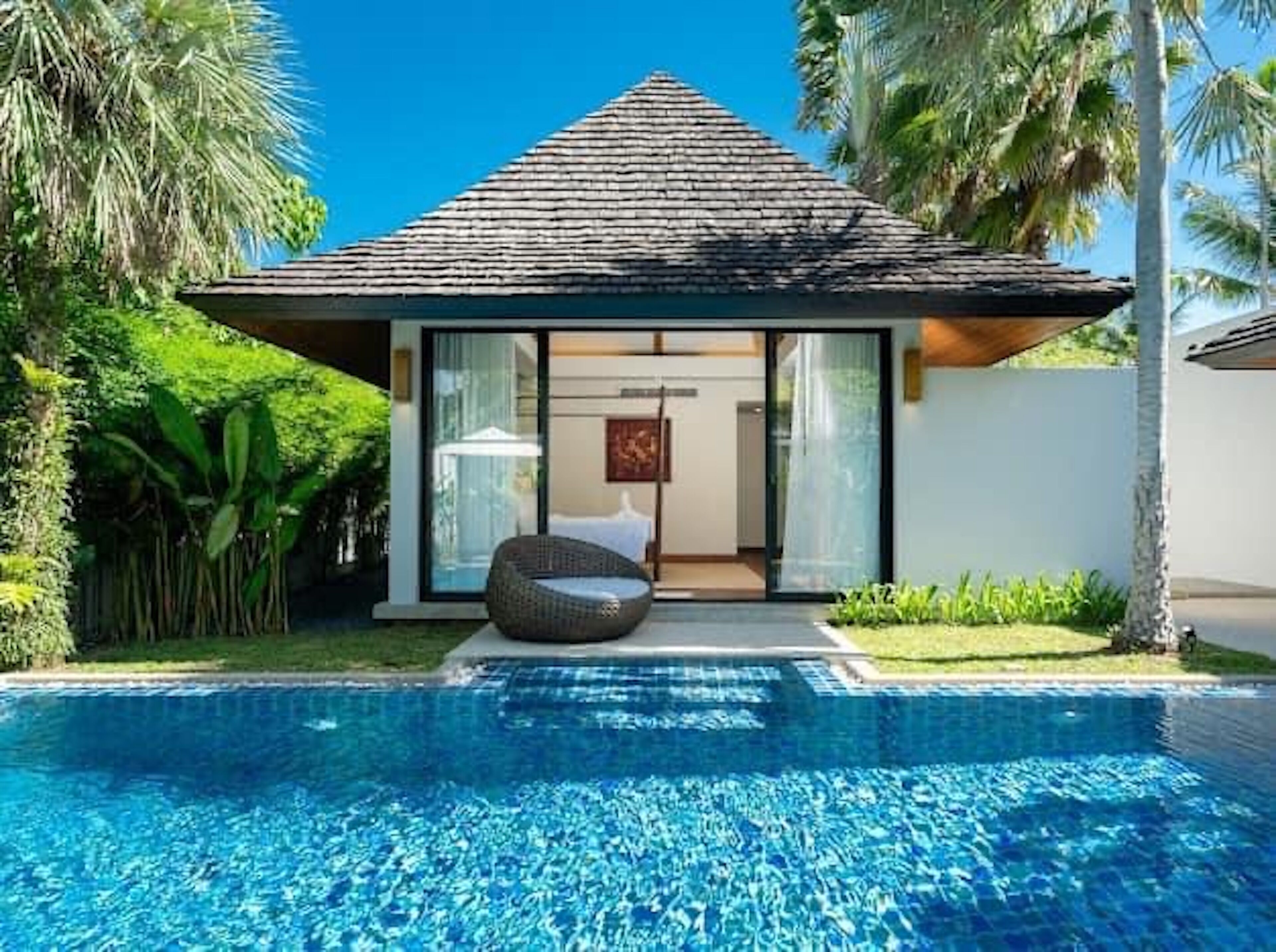 A modern villa with a swimming pool, a covered outdoor seating area, and lush tropical vegetation.