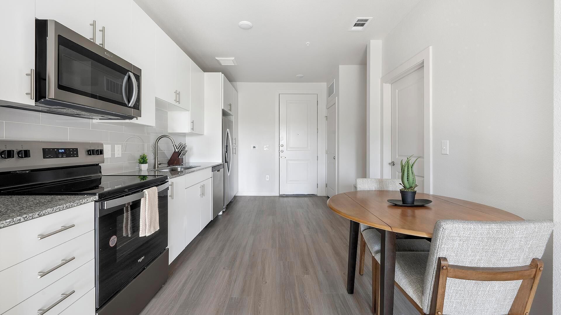 Une cuisine moderne avec des meubles blancs, une cuisinière noire et un four à micro-ondes. On y trouve un coin repas avec une table et des chaises en bois, ainsi qu’une plante en pot sur la table.