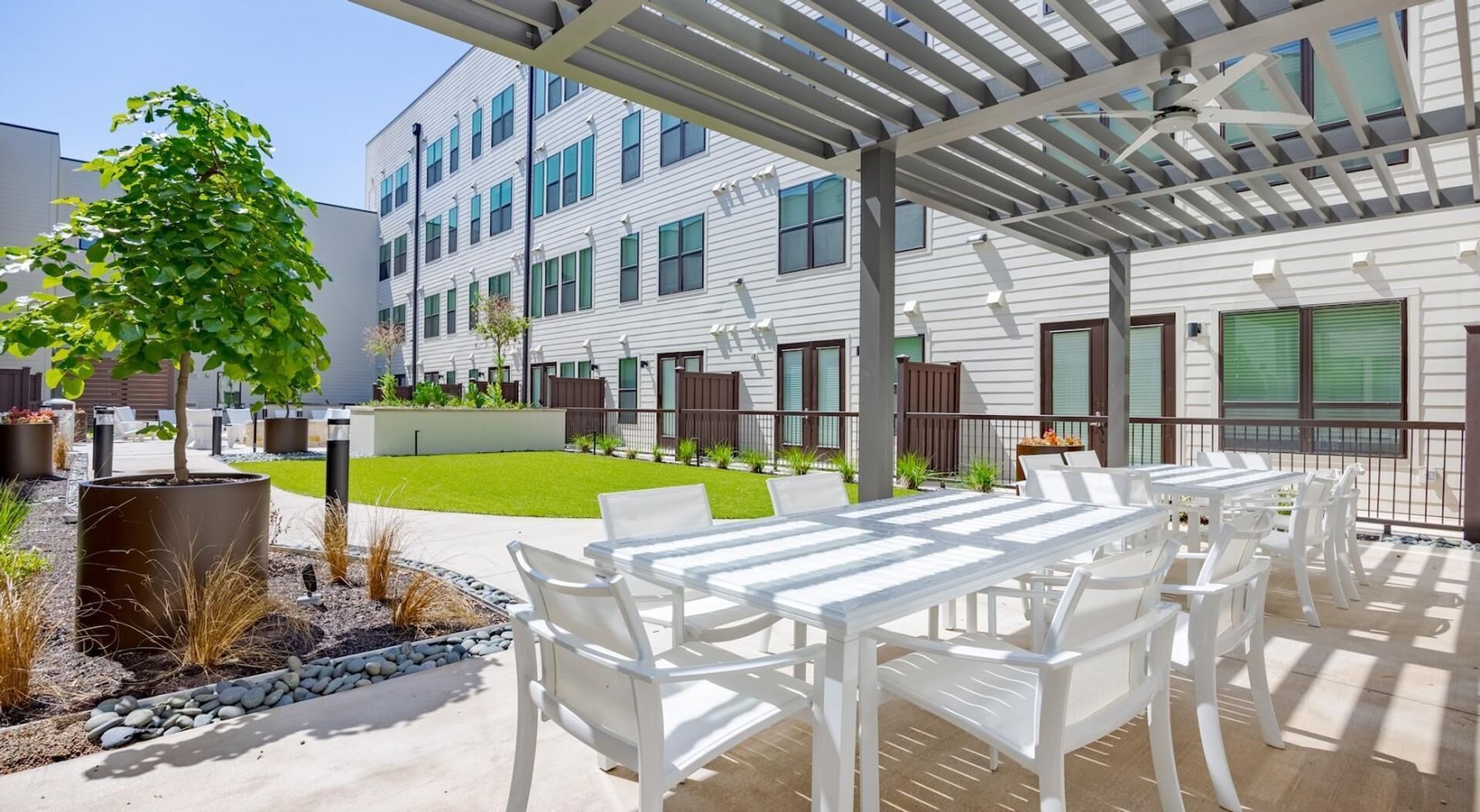 A covered outdoor patio with white furniture, a small lawn, and a building with multiple windows in the background.