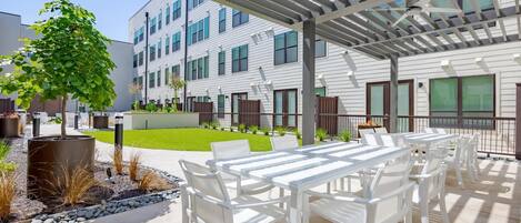 A covered outdoor patio with white furniture, a small lawn, and a building with multiple windows in the background.