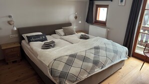 Ein ordentlich bezogenes Bett mit einer Karo-Decke, einer Nachttischlampe, einem kleinen Holennachttisch und einem Fenster mit Vorhängen.