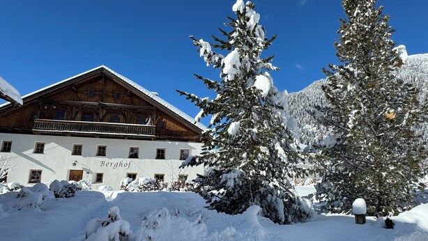 Ein traditionelles Holzhaus mit einem Schild, auf dem "Berghof" steht, umgeben von schneebedeckten Bäumen und blauem Himmel.