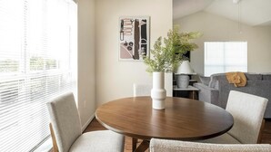Ett modernt vardagsrum med ett runt träbord, två stolar, en grå soffa, en vit vas med gröna växter och ett inramat, abstrakt konstverk på väggen.