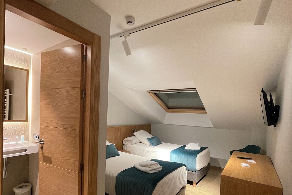 Et moderne hotelværelse med en trædør, ovenlys, en stor seng med et grønt tæppe, et natbord og et lille skrivebord.