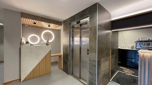 Un espace d’accueil moderne avec un comptoir de réception en bois, un ascenseur en verre et un coin café.