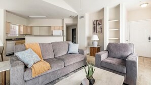 Een moderne woonkamer met een grijze bank, een bijpassende fauteuil en een salontafel. Achtergrond toont een keuken met witte kasten en een raam met zonwering.