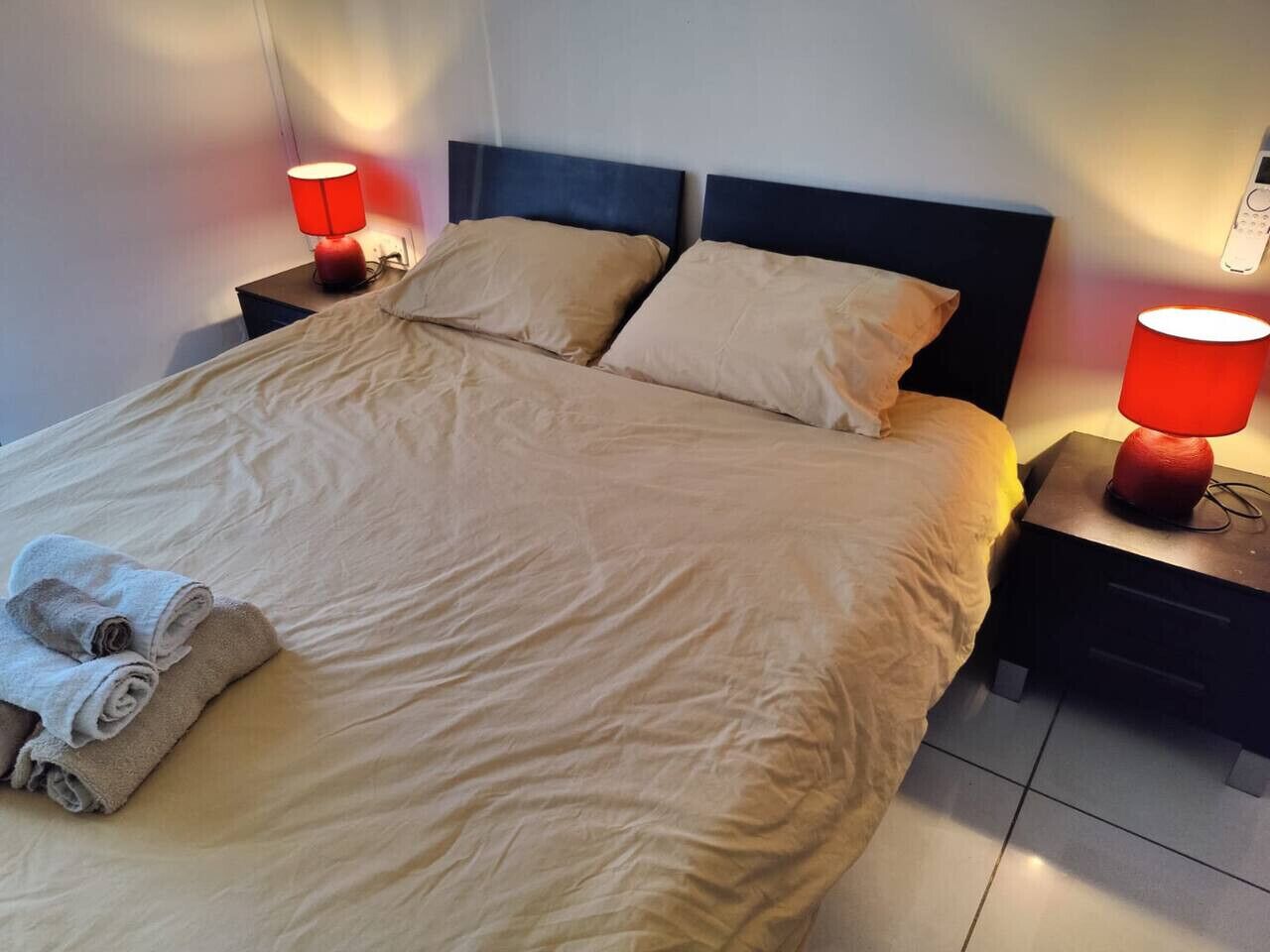 Un lit bien fait, avec du linge de lit beige, deux oreillers et une pile de serviettes roulées sur le côté gauche. De chaque côté du lit, on trouve deux tables de chevet sur lesquelles sont posées des lampes rouges.