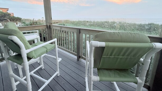 A wooden deck with two white chairs and a table, overlooking a beach with tall grass and a clear sky. - Anchor & Dreams - Peaceful Updated 2BR Oceanfront Condo (Ocean Isle Beach)