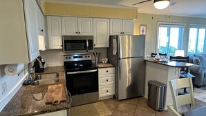 A modern kitchen with white cabinets, stainless steel appliances, a granite countertop, and a dining area with a view. - Anchor & Dreams - Peaceful Updated 2BR Oceanfront Condo (Ocean Isle Beach)