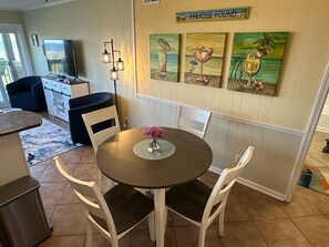A dining area with a round table, white chairs, and a vase of flowers. There are three paintings on the wall above the table, and a sign that reads "PARADISE FOUND". - Anchor & Dreams - Peaceful Updated 2BR Oceanfront Condo (Ocean Isle Beach)