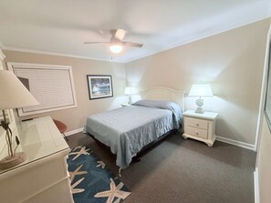 A bedroom with a bed, bedside lamps, a ceiling fan, a window with blinds, and a framed picture on the wall. - Anchor & Dreams - Peaceful Updated 2BR Oceanfront Condo (Ocean Isle Beach)