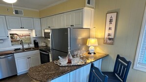 A kitchen with white cabinets, a granite countertop, a refrigerator, a sink, and a dining area with blue chairs. - Anchor & Dreams - Peaceful Updated 2BR Oceanfront Condo (Ocean Isle Beach)