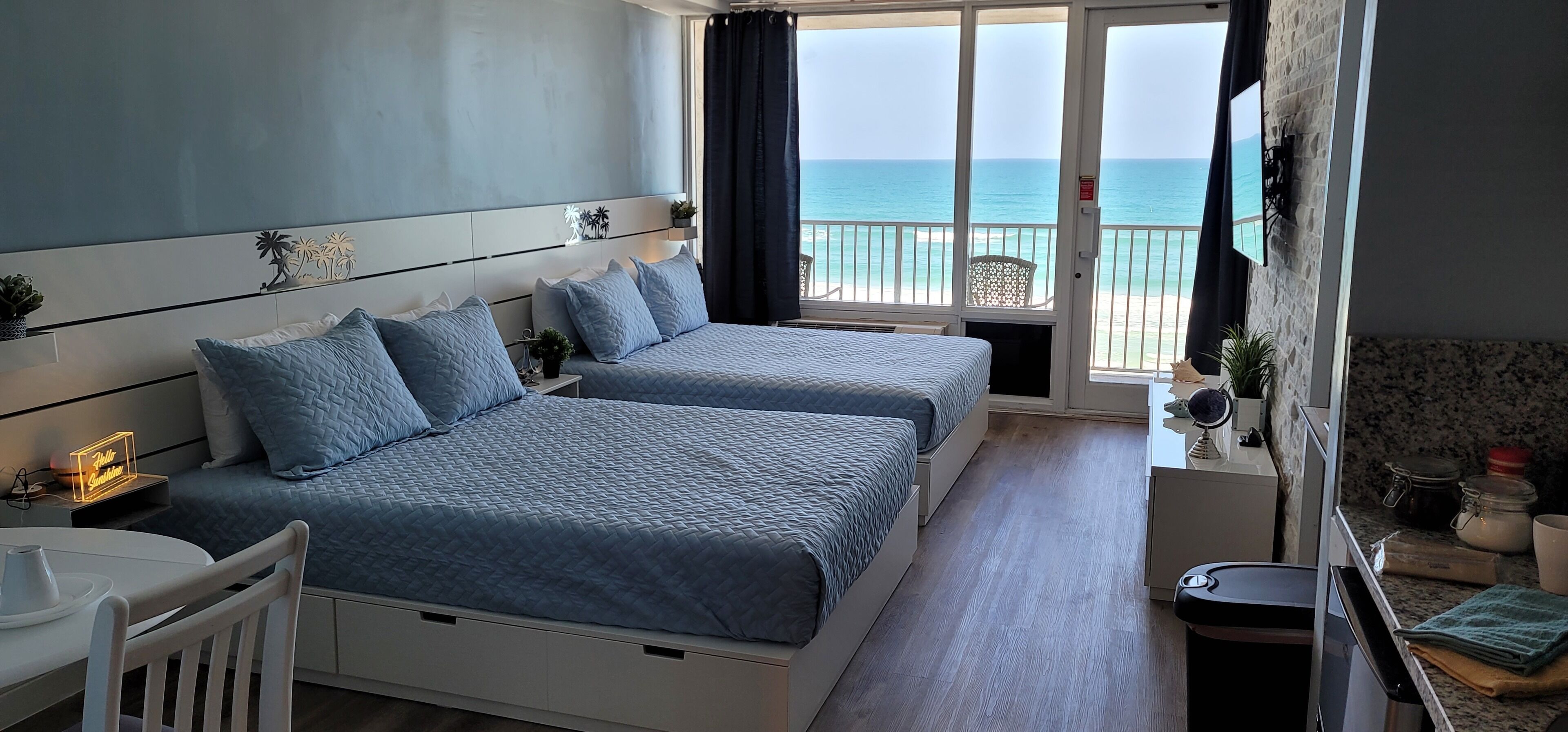 A hotel room with two beds, a balcony with a view of the sea, a small table with a chair, and a kitchenette area.
