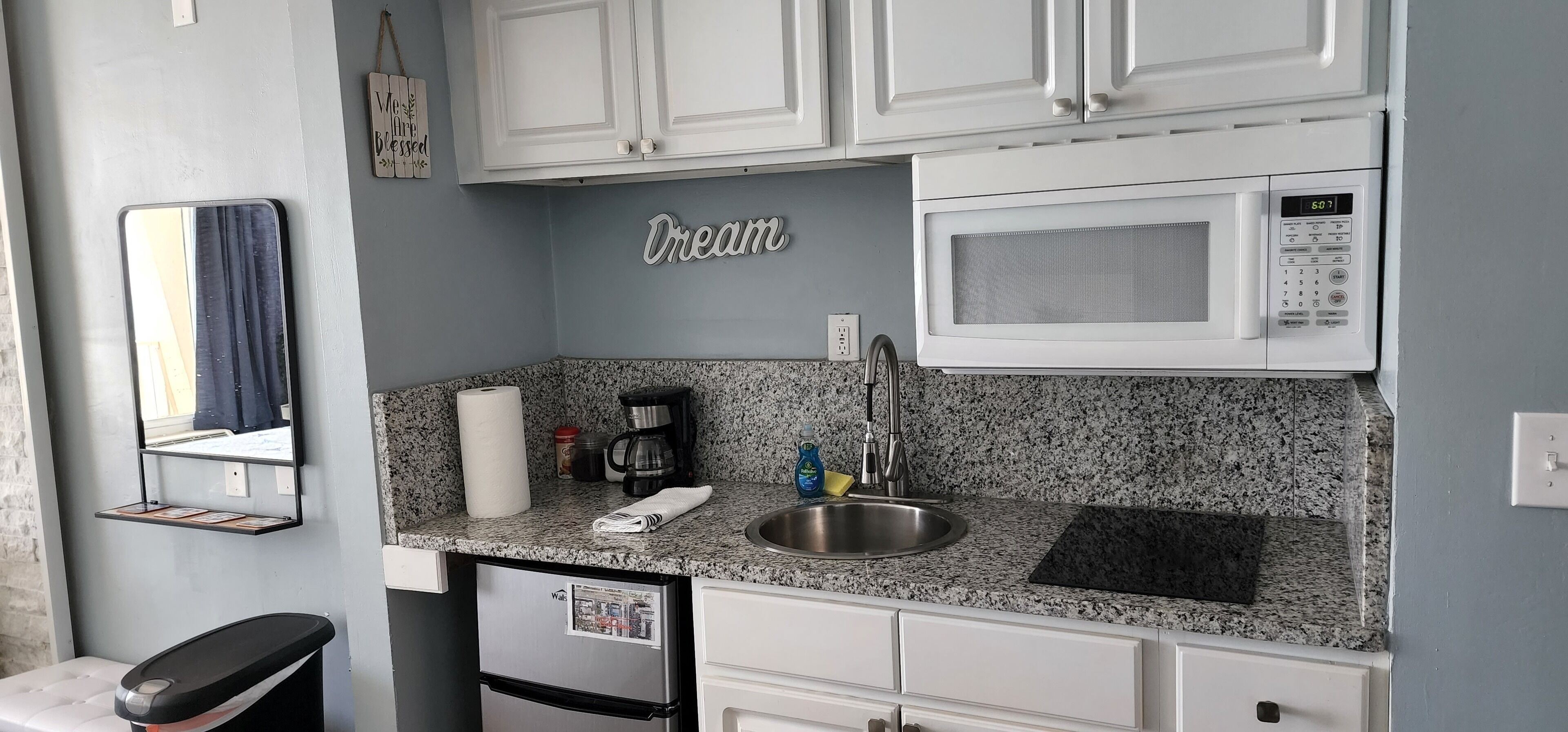 A compact kitchenette with white cabinets, a microwave, a sink, and a countertop with a paper towel holder, coffee maker, and cleaning supplies.