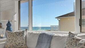 A white sofa with patterned cushions and a blue throw blanket, positioned by a window overlooking a beach and a building. - Updated End Unit in Wrightsville Dunes with Loft and Oceanfront Views (Wrightsville Beach)