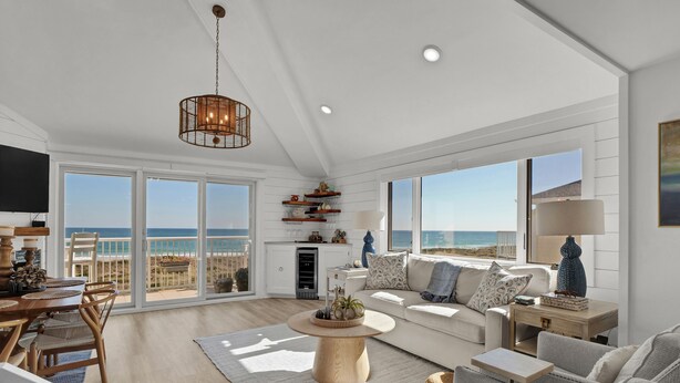 A spacious living room with a large window overlooking the beach, a wooden coffee table, and a cozy seating area. - Updated End Unit in Wrightsville Dunes with Loft and Oceanfront Views (Wrightsville Beach)