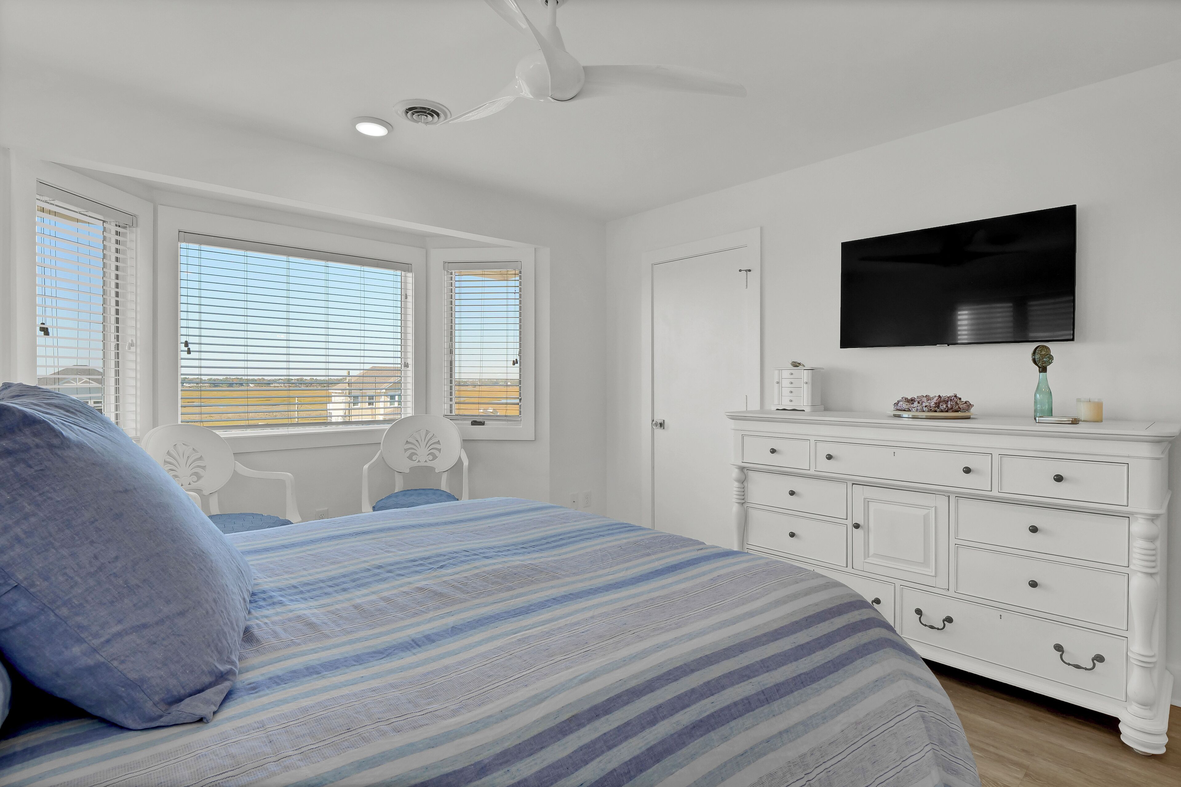 A bedroom with a large bed, a white dresser, a flat-screen TV, and two windows with blinds.
