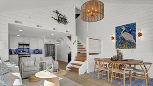 A modern living room with a sofa, coffee table, and dining area with wooden chairs and a staircase. - Updated End Unit in Wrightsville Dunes with Loft and Oceanfront Views (Wrightsville Beach)