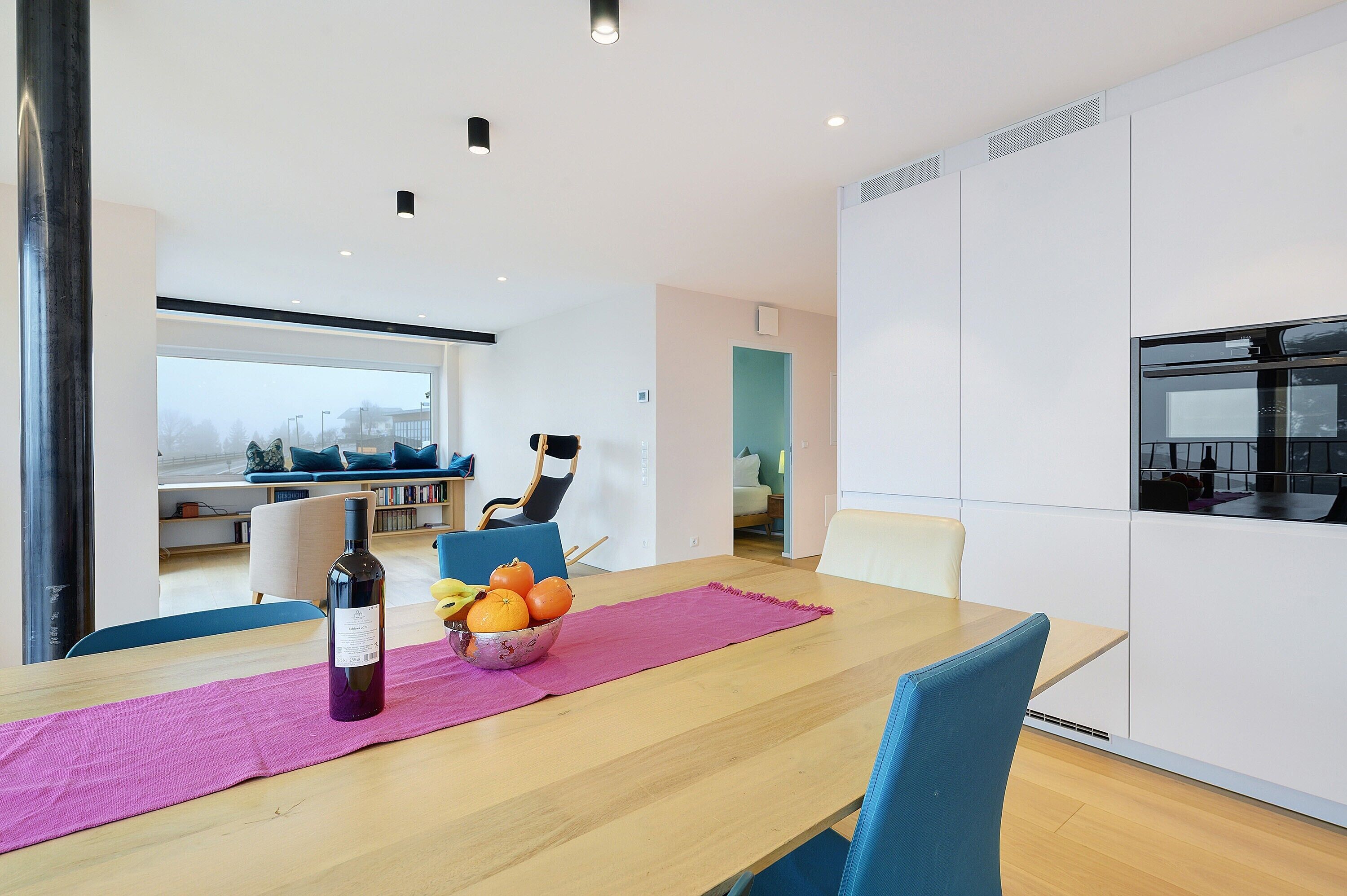 A modern dining area with a wooden table, a pink placemat, a bowl of fruit, and a bottle of wine.