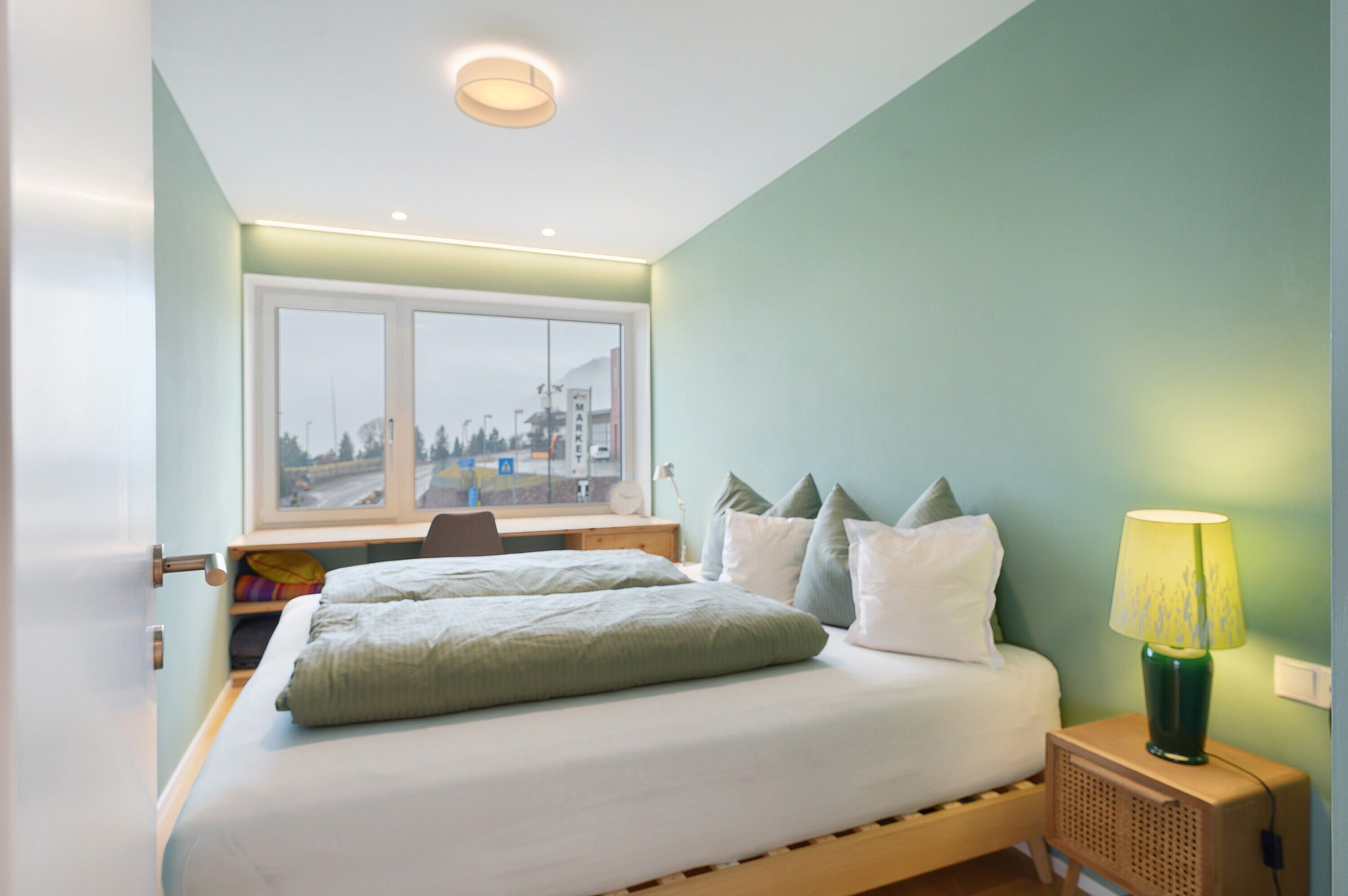 Een slaapkamer met een bed, kussens, een bureau, een lamp en een raam met uitzicht op gebouwen.
