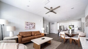 A modern living room with a brown sofa, a coffee table, and a dining area with a wooden table and chairs. There is a kitchen in the background with white cabinets and stainless steel appliances. - Landing | Beautiful 2BD, Gym, Pool (Palm Bay)