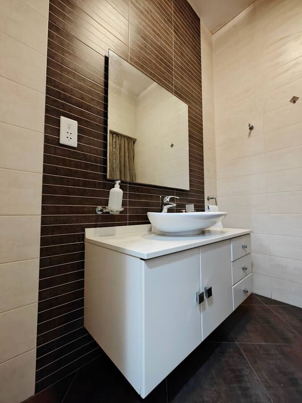 Une salle de bain moderne avec un meuble-lavabo blanc, des murs recouverts de carreaux foncés, un grand miroir et un lavabo blanc.