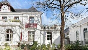 Ein weißes Gebäude mit einem Uhrturm, einem Balkon mit roten Geländern und einem Baum im Vordergrund.
