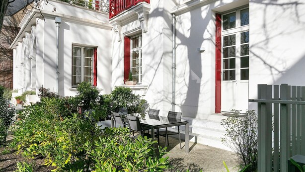 Ein weißes Gebäude mit roten Fensterläden, einem Balkon und einer Terrasse mit Gartenmöbeln.