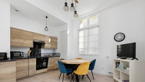 Eine moderne Küche mit Holzschränken, einem Esstisch mit blau und gelben Stühlen, einem Fernseher auf einem weißen Schrank und einer Uhr an der Wand.