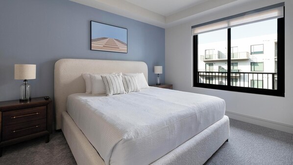 A bedroom with a large bed, two bedside tables with lamps, a framed picture on the wall, and a window with a view of buildings. - Landing | Beautiful 2BD, Pool, Tennis Court (Houston)