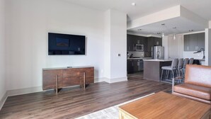 A modern living room with a wooden floor, a brown leather sofa, a wooden coffee table, and a flat-screen TV mounted on the wall. - Landing | Spectacular 2BD, Gym, Pool (Houston)