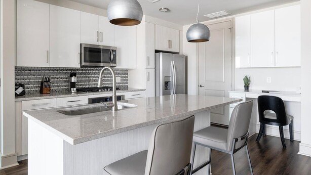 A modern kitchen with white cabinetry, a central island with bar stools, stainless steel appliances, and pendant lighting. - Landing | Incredible 2BD, Tennis Court, Pool (Houston)