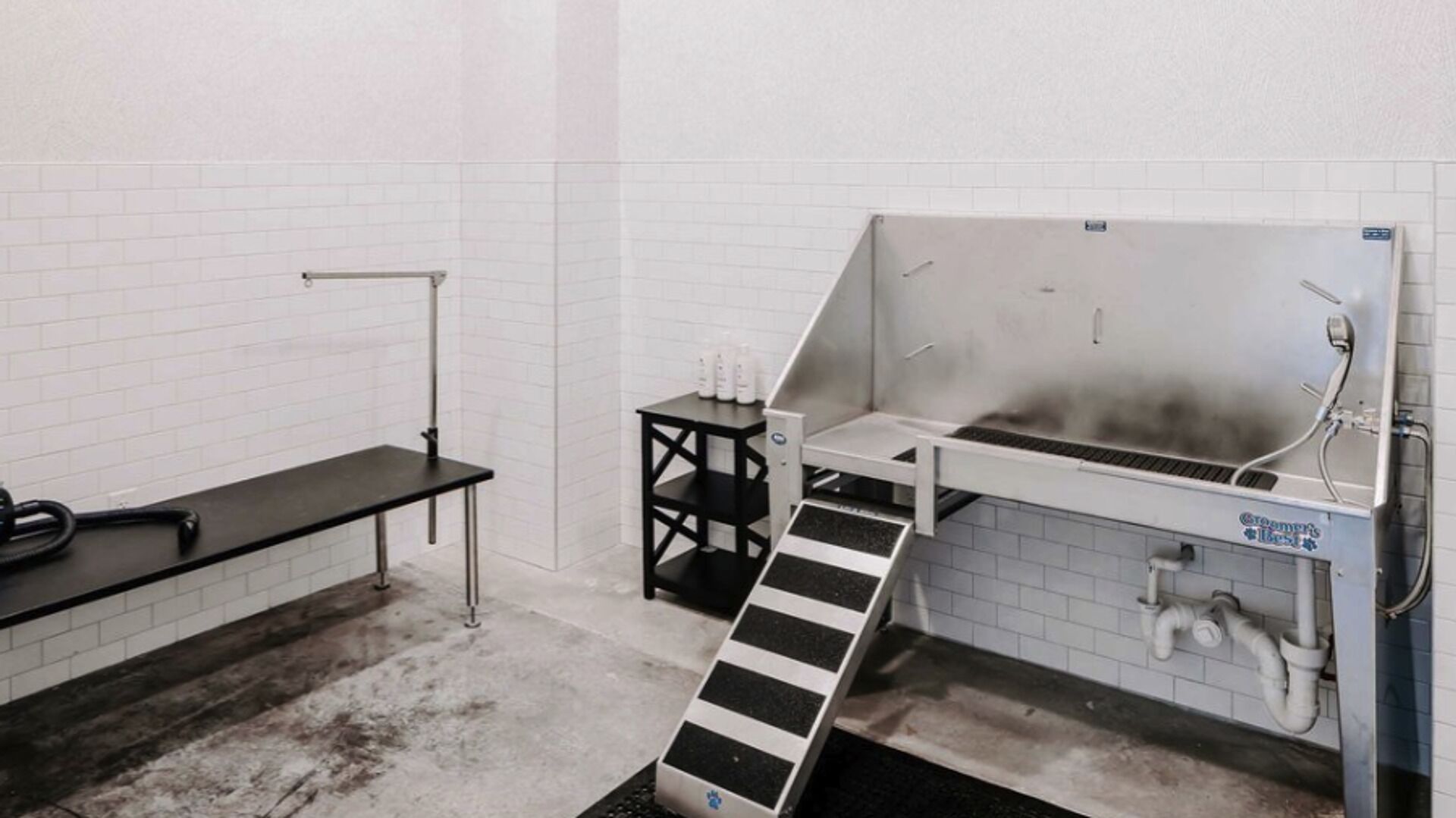Een badkamer met een witte betegelde muur, een zwarte wastafel, een klein zwart tafeltje met toiletartikelen en een douchegedeelte met een metalen ladder.