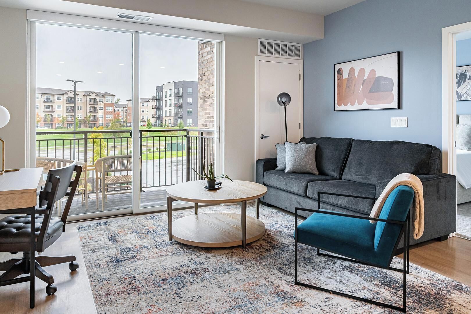 Un salón moderno con un sofá gris, un sillón azul, una mesa de centro redonda y vistas a un balcón con barandilla.