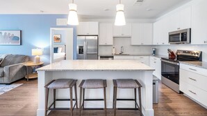 A modern kitchen with white cabinetry, a central island with bar stools, stainless steel appliances, and pendant lighting. - Landing | Amazing 2BD, Pool, Clubhouse (Vero Beach)