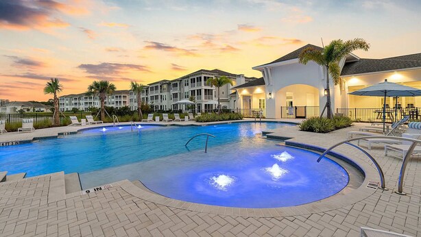A swimming pool with a 9-inch depth marker, surrounded by lounge chairs and palm trees, with a backdrop of multi-story buildings and a sunset sky. - Landing | Amazing 2BD, Pool, Clubhouse (Vero Beach)