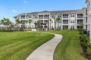 A modern multi-story residential building with a central courtyard, a curved pathway, and surrounding greenery. - Landing | Incredible 2BD, Clubhouse, Gym (Vero Beach)