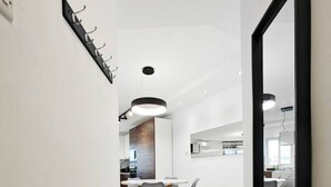 A modern, well-lit hallway with a dining area in the background, featuring a large mirror and a metal rack. - Valley view 2  2 bedroom aparment (Luqa)