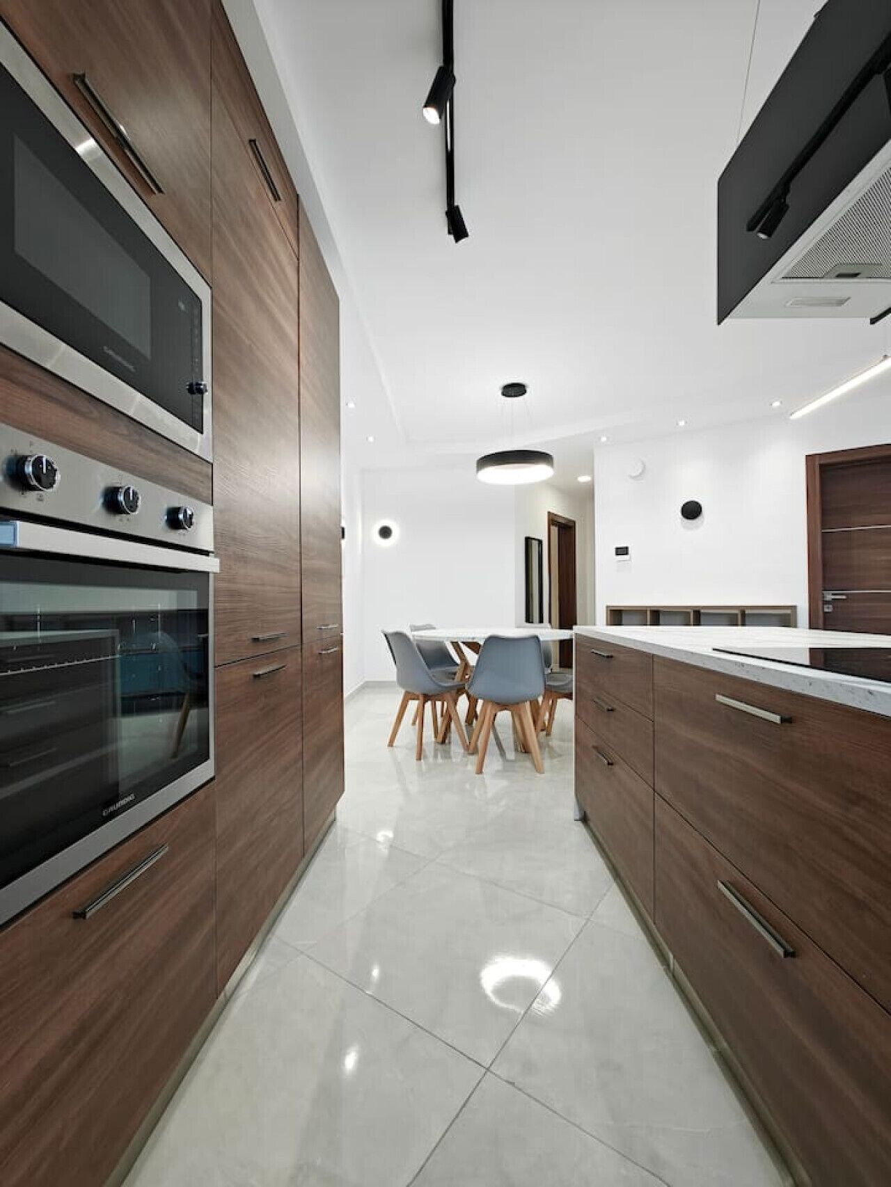 Een moderne keuken met inbouwovens, een eethoek met tafel en stoelen, en een strakke, witte betegelde vloer.